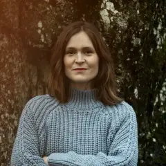 Elin Cullhed, klädd i en ljusblå stickad tröja, står med armarna i kors lutad mot ett mossigt träd.