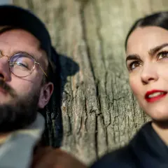 En man med glasögon och skägg lutar sig mot ett träd, bredvid sin mika partner.