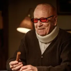 Äldre man med nackkrage och röda glasögon håller en cigarr. Han ser ut som grannen i beck, redo för en stunds avkoppling.