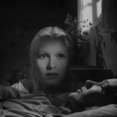 De två kvinnorna i dramatisk belysning. En kvinna ligger ner, medan den andra tittar ner på henne med intensiv blick.
