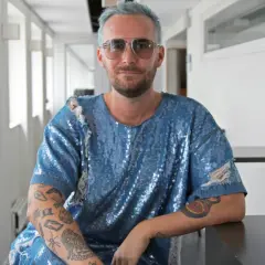 Fredrik Robertsson, med blått hår och solglasögon, sitter i en glittrig blå tröja med slitna detaljer. Hans armar är tatuerade.