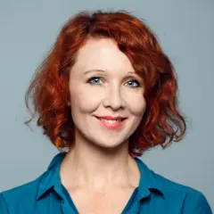 Ellinor Skagegård, en kvinna med rött lockigt hår och blå ögon, ler mot kameran i en blå skjorta.