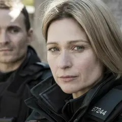 Två poliser i uniform, en kvinna i förgrunden med intensiv blick, bakgrunden svagt belyst av blåa ljus.