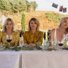 Fem kvinnor, kanske **bad sisters**, sitter vid ett dukat bord utomhus. En bär ögonlapp, en annan en glittrig gul klänning.