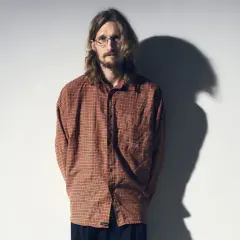 Marcus Berggren komiker, med sitt karaktäristiska hår och glasögon, står framför en vit vägg med en skugga som dansar bakom honom.