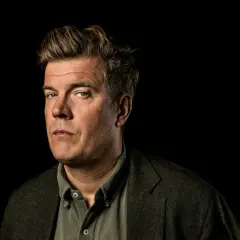 Gunnar Ardelius, en man med intensiv blick och rufsig frisyr, bär en tweedkavaj över en skjorta.