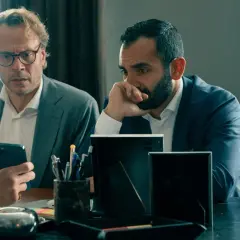 Två män i kostym sitter vid ett bord. Den ena mannen, med glasögon, tittar på sin mobiltelefon. Den andra mannen, med skägg, funderar djupt. Scen från "hundarna" på SVT.
