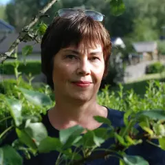 Anna Bratt, en kvinna med glasögon på huvudet, ler mot kameran i en lummig trädgård.