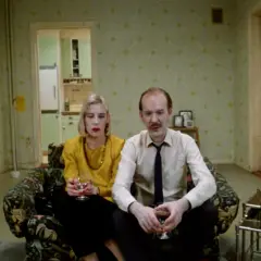 Ett par sitter i en soffa, en kvinna med gul tröja och en man i skjorta och slips. Kanske en scen från en Roy Andersson reklamfilm.