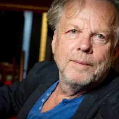 En äldre man med grått hår och skägg, klädd i kavaj och blå tröja. Han påminner om en ung Wallander.