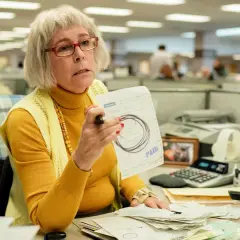 Jamie Lee Curtis som en kontorist i filmer, med glasögon och en gul polotröja, håller upp ett papper med texten "PAID".