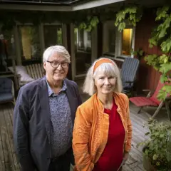 Ett par står på en altan. Mannen har glasögon och en blå kavaj. Kvinnan har ett orange hårband och en orange jacka. De ser ut som en del av amanda romare familj.