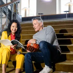 En kvinna i gul dräkt läser en bok medan en man med basket håller ett samtal. Kanske de diskuterar hur man kan jobba med att skriva om sport.