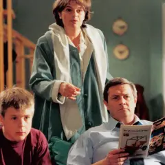Familjen Svensson samlad i vardagsrummet. Pappa läser tidningen, mamma står och pratar, sonen sitter bredvid.