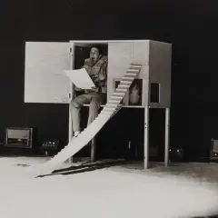 En man sitter i en träkonstruktion och läser högt ur ett papper. Robert Rauschenberg kan ha inspirerat denna scen med sina experimentella verk.