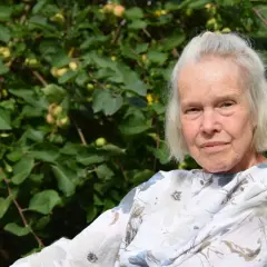 Äldre kvinna med grått hår ler mot kameran, bakom henne syns ett äppelträd. Merete Mazzarella njuter av sommaren.