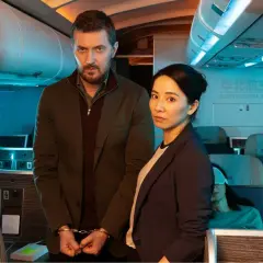 Man med handbojor och kvinna står i ett flygplan. En sovande passagerare syns i bakgrunden. Scen från konspirationen serie.
