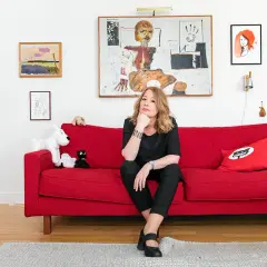 Åsa Linderborg sitter på en röd soffa, omgiven av konst och leksaker som påminner om barn.