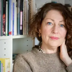 Elisabeth Åsbrink, med blå ögon och lockigt hår, sitter framför en bokhylla. Hon verkar lite sjuk, men hennes blick är klar.
