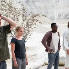 Fyra personer står i en öde, ljus miljö. En man med händerna i håret, en kvinna med blicken fäst framåt, en man med öppen mun och en man i vit, gammaldags klädsel. Scen från filmen Midsommar.
