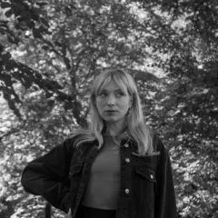 Sandra Beijer, med sitt blonda hår och blick mot horisonten, står i en skog. Hon bär en jeansjacka.