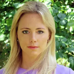 Close-up portrait of a blonde woman with green eyes and a purple top, against a blurred green foliage background.