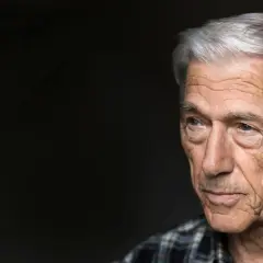 Porträtt av en äldre man med grått hår och rynkor, Theodor Kallifatides, som blickar åt sidan mot en mörk bakgrund.