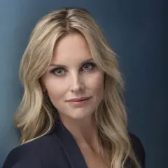 Porträtt av Emelie Schepp med blont hår och blå ögon, klädd i en mörk kavaj mot en blå bakgrund.