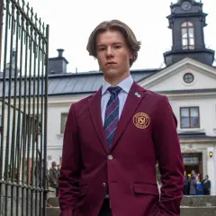 En ung man i skoluniform, som påminner om unga kungligheter, står framför en ståtlig byggnad.