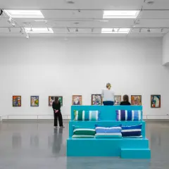 I en konsthall sitter besökare på en turkos scen med kuddar, omgivna av färgglada tavlor. Kanske är de där för att inspireras, för att vara en musa för konstnären.