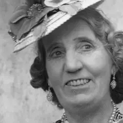 Julia Cæsar, en glad dam med hatt och örhängen, ler mot betraktaren.