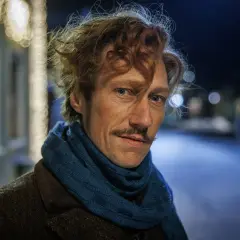 Man med rufsigt hår och mustasch, klädd i brun rock och blå halsduk. Kanske en del av rollistan i jakten på en hemlighet i vintermörkret.