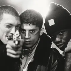 Tre unga män, en med en pistol riktad mot kameran, i en dramatisk scen från "La Haine" (Medan vi faller).