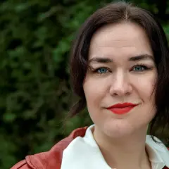 Camilla Sten med blå ögon och rött läppstift, klädd i en röd jacka, ler mot kameran.