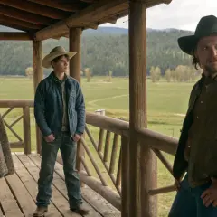 Tre personer på en veranda med berg i bakgrunden. En kvinna med mönstrad kofta, en ung man med cowboyhatt och en man med hatt och brun jacka. Scen från Yellowstone-serien.