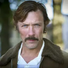 En manlig svensk skådespelare med blå ögon och mustasch, klädd i en brun jacka med foder.