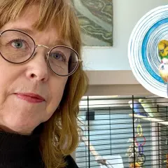 Aase Berg med glasögon tittar mot kameran, med en fågelbur och en teckning av två undulater i bakgrunden.