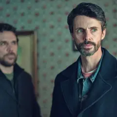Två män i mörka rockar står inomhus. Den främre mannen har intensiv blick, den bakre är suddig. Kanske är de från dept q.