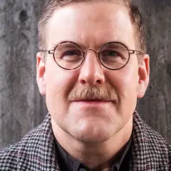 Adam Svanell med glasögon och mustasch, klädd i en rutig rock, framför en grå bakgrund.
