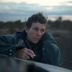 En kvinna med en cigarett i handen lutar sig mot en bil i ett öde landskap. Hon är en del av rollistan i Nomadland.
