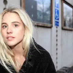En ung kvinna med blont hår, Paolo Nobis Sandén, tittar in i kameran framför en grå byggnad med fönster.