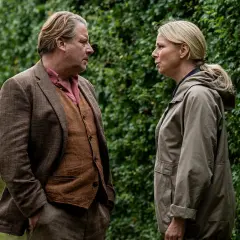 Två personer står vända mot varandra framför en grön häck. Mannen bär kavaj och väst, kvinnan en jacka. Scen från en dansk kriminalserie.
