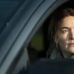 En kvinna med intensiv blick sitter i en bil, kanske på väg att lösa ett fall som i Mare of Easttown.