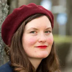 Alva Dahl, med en vinröd basker och röda läppar, ler mot kameran. Hon står utomhus vid ett träd.