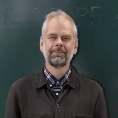 Anders, med grått hår och skägg, ler mot kameran framför en grön tavla. Han bär en rutig skjorta under en mörk jacka.
