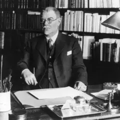 Albert Engström, med glasögon och kostym, sitter vid sitt skrivbord med böcker i bakgrunden. Penslar och bläckhorn syns på bordet.