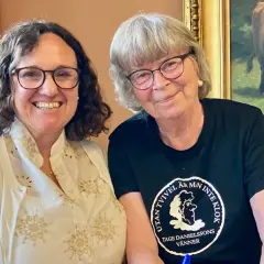 Maria Martinsson och en vän ler mot kameran. Bakom dem hänger en tavla med en ko.