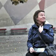 Elisabeth Rynell sitter på en bänk med en bok i knät, blickar ut mot en geometrisk vägg.