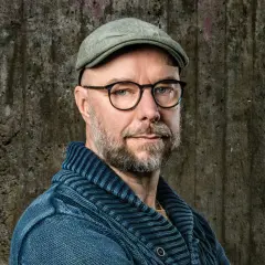 Sven Olov Karlsson, en man med glasögon och keps, blickar mot kameran. Han bär en blå stickad tröja.