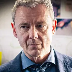 En man i kostym, med grånande hår, blickar fundersamt framåt. Han är en del av grace serie.